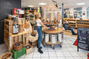 A young customer stands in front of a shelf with filling containers for muesli in an unpacked shop. A boy is swinging to the right. Scene from an unwrapped shop. A young customer stands in front of a shelf with filling containers for muesli in an unpacked shop. A boy is swinging to the right. Scene from an unwrapped shop.